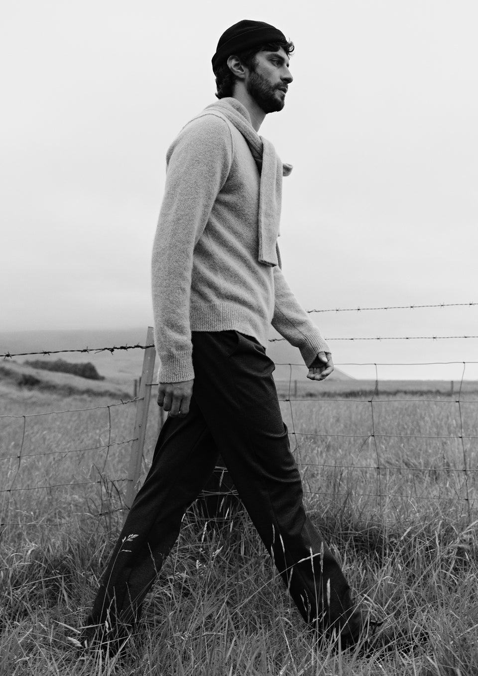 A man in a light gray knit sweater and dark trousers walks through a grassy field near a wire fence, shown in a black and white photograph.