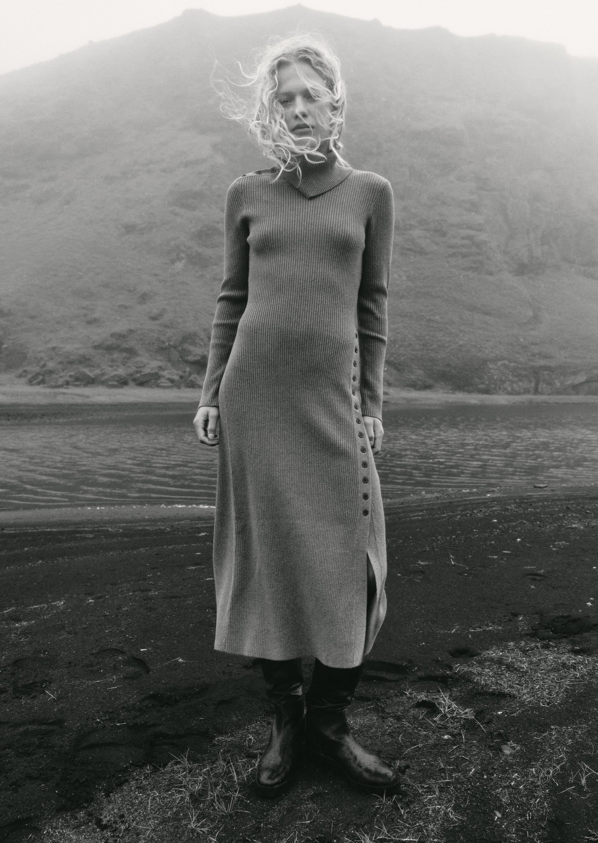 A woman stands outdoors on a dark shore in front of water and a mountain, wearing a long fitted knit dress with side buttons and dark boots.