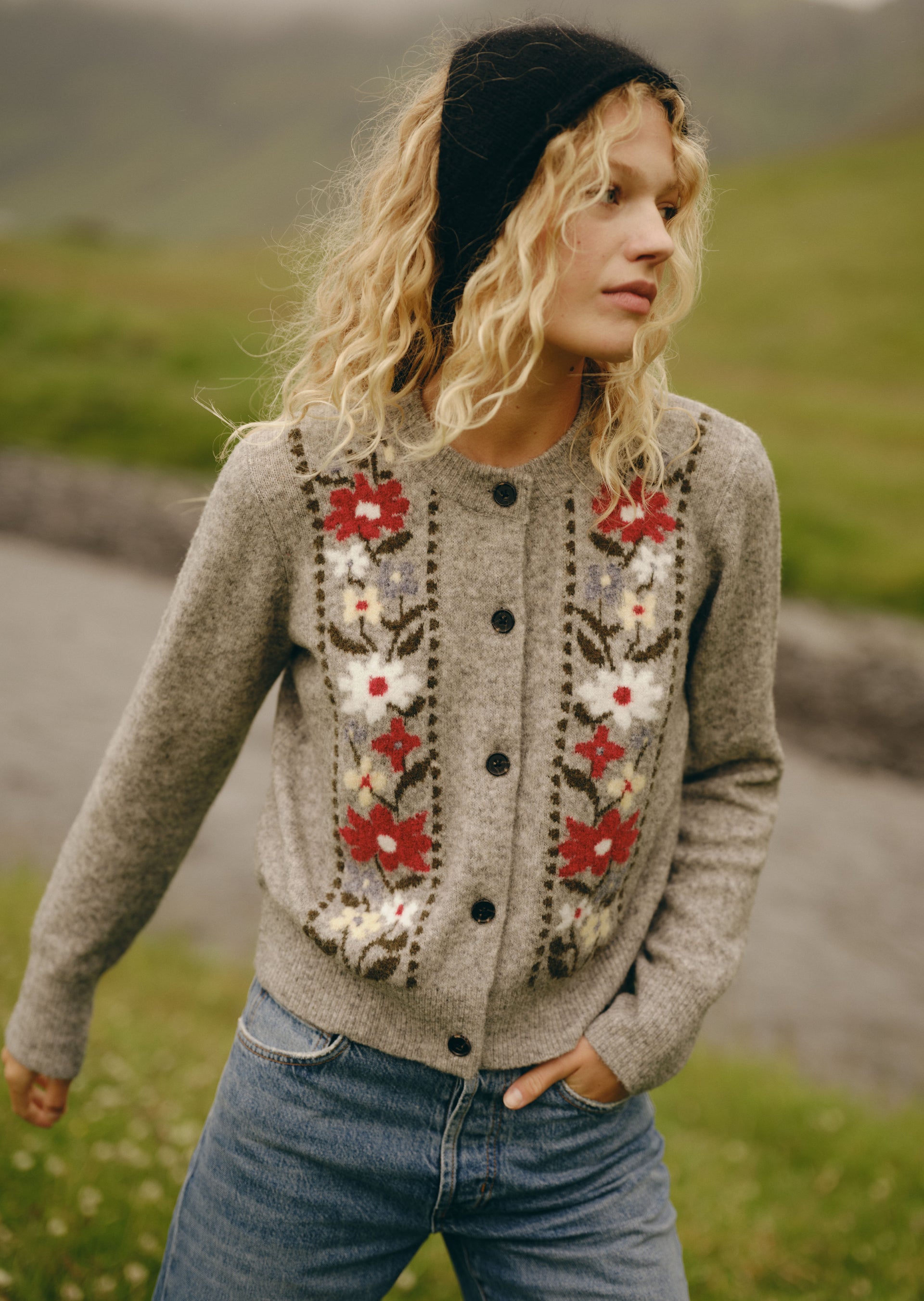 A woman wearing a gray knit cardigan with red and white floral embroidery, blue jeans, and a black beanie, standing outdoors in a grassy field.
