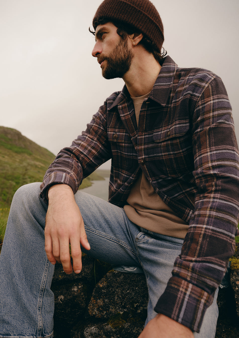 A man wearing a brown knit beanie, a dark brown and black plaid flannel shirt over a tan t-shirt, and blue jeans, sitting outdoors on a rock with a hazy, overcast sky and green hills in the background.