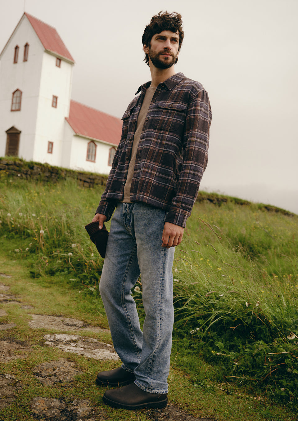 A man wearing a dark brown and black plaid flannel shirt over a beige t-shirt, blue jeans, and dark brown boots, standing outdoors on a grassy hill with a white church in the background.
