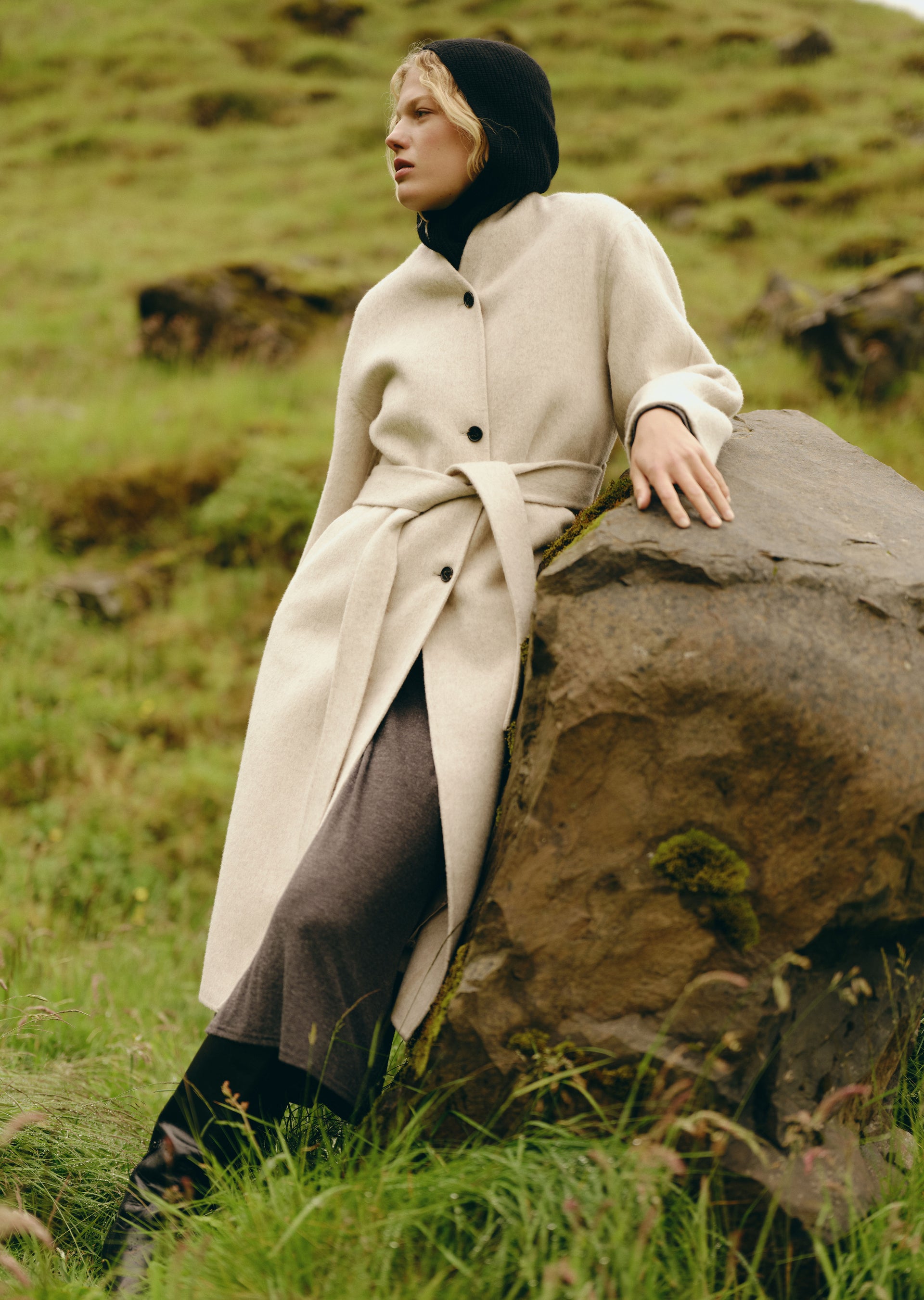 A woman wearing a long cream wool coat with a tie belt, a black head covering, and dark trousers, posing outdoors on a large mossy rock in a grassy field.