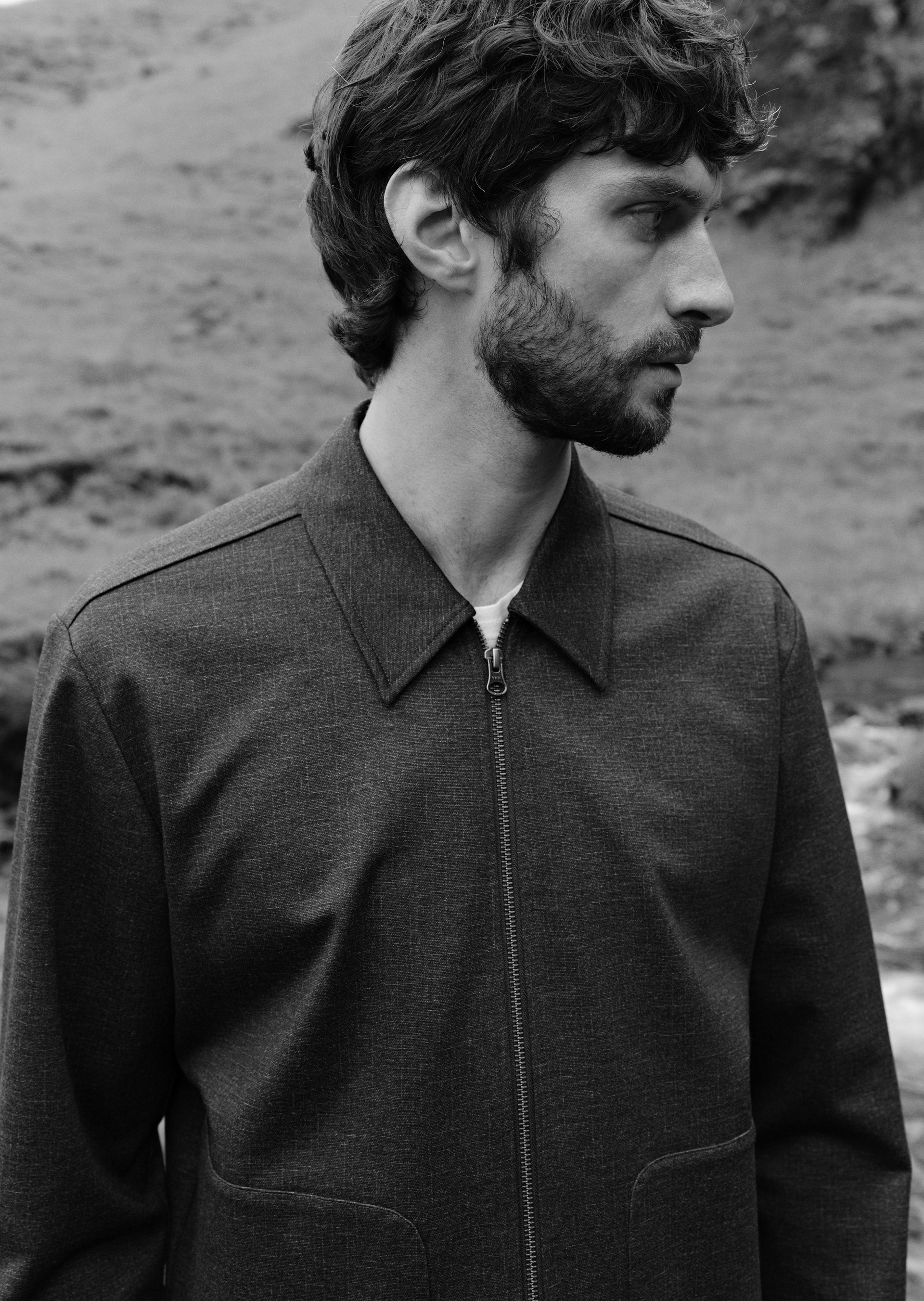 A man in profile wearing a dark textured zip-front jacket, photographed outdoors against a blurred natural background. The image is in black and white.