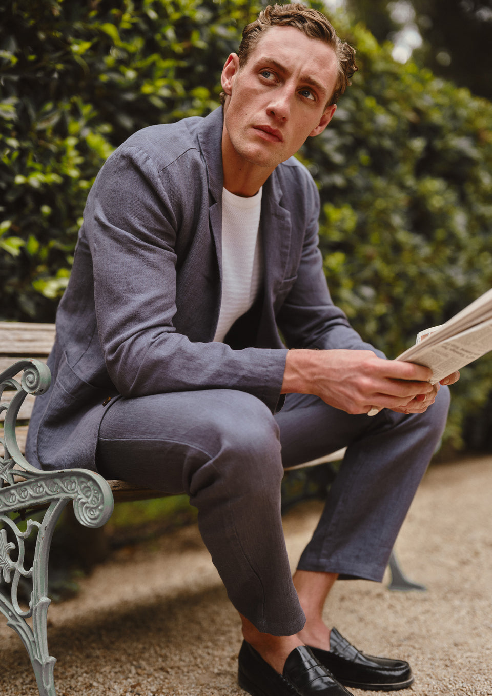 Man in a blue suit sitting on a bench reading a newspaper with greenery in the background