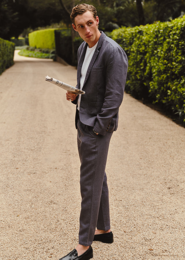 Man in a blue suit holding a newspaper on a path with greenery.
