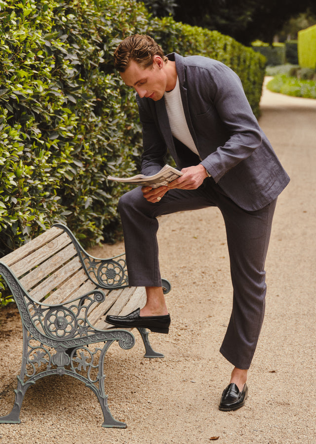 Man in a suit reading a newspaper on a bench in a park.