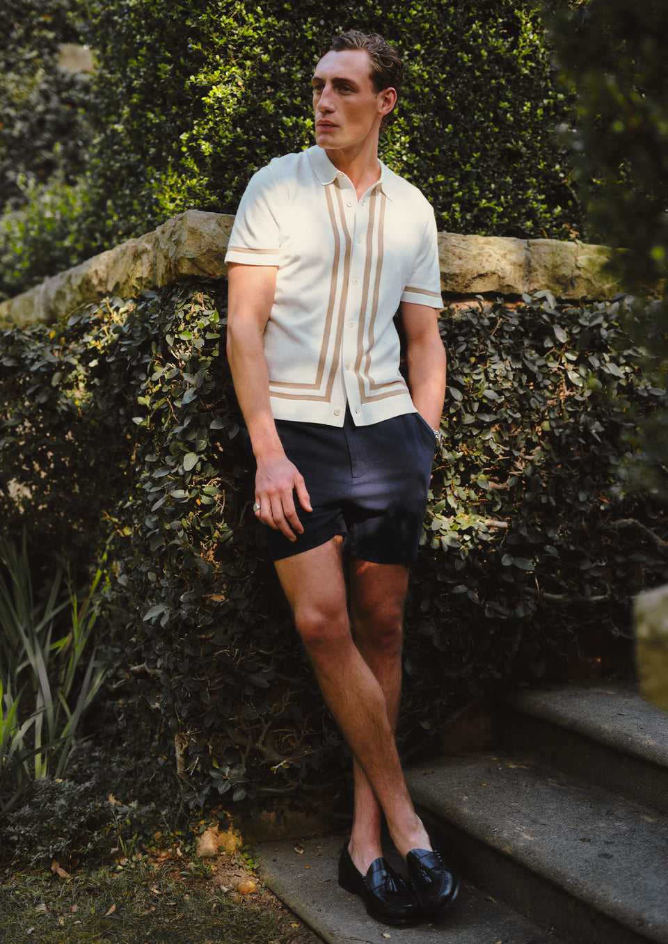 Man in casual attire standing outdoors against a stone wall with greenery.
