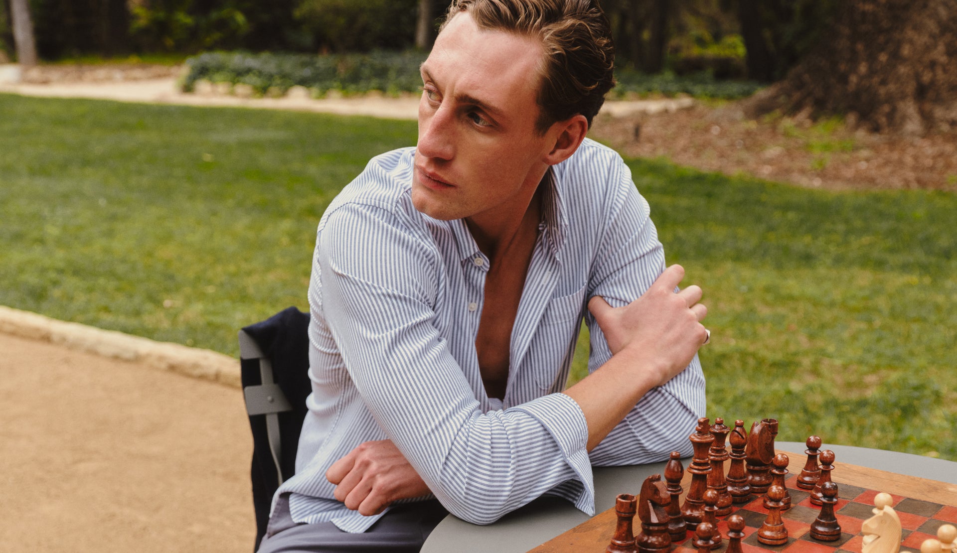 Man sitting at a chessboard outdoors with trees and grass in the background