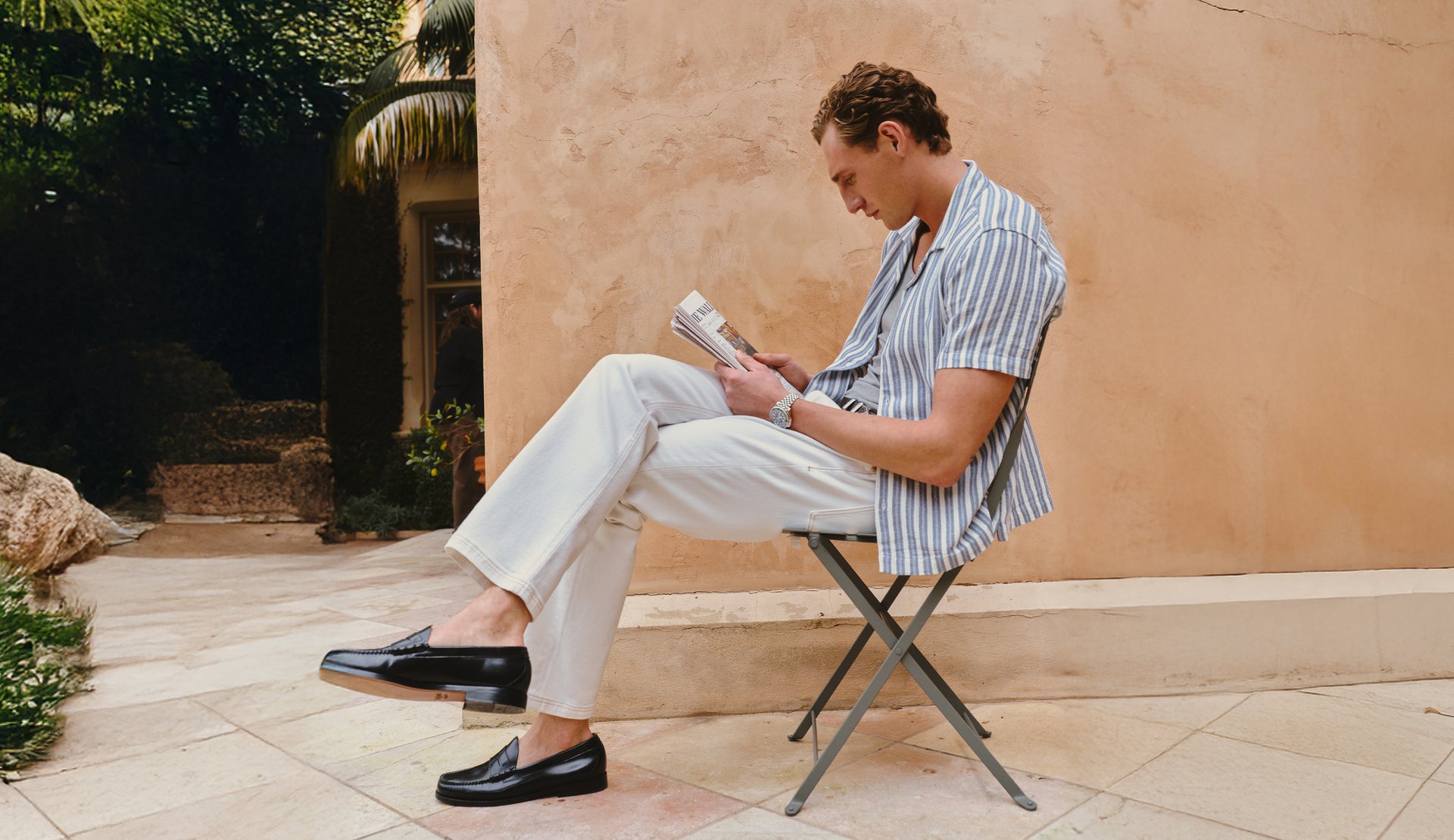 Man sitting on a chair reading a newspaper outdoors with a beige wall and greenery in the background
