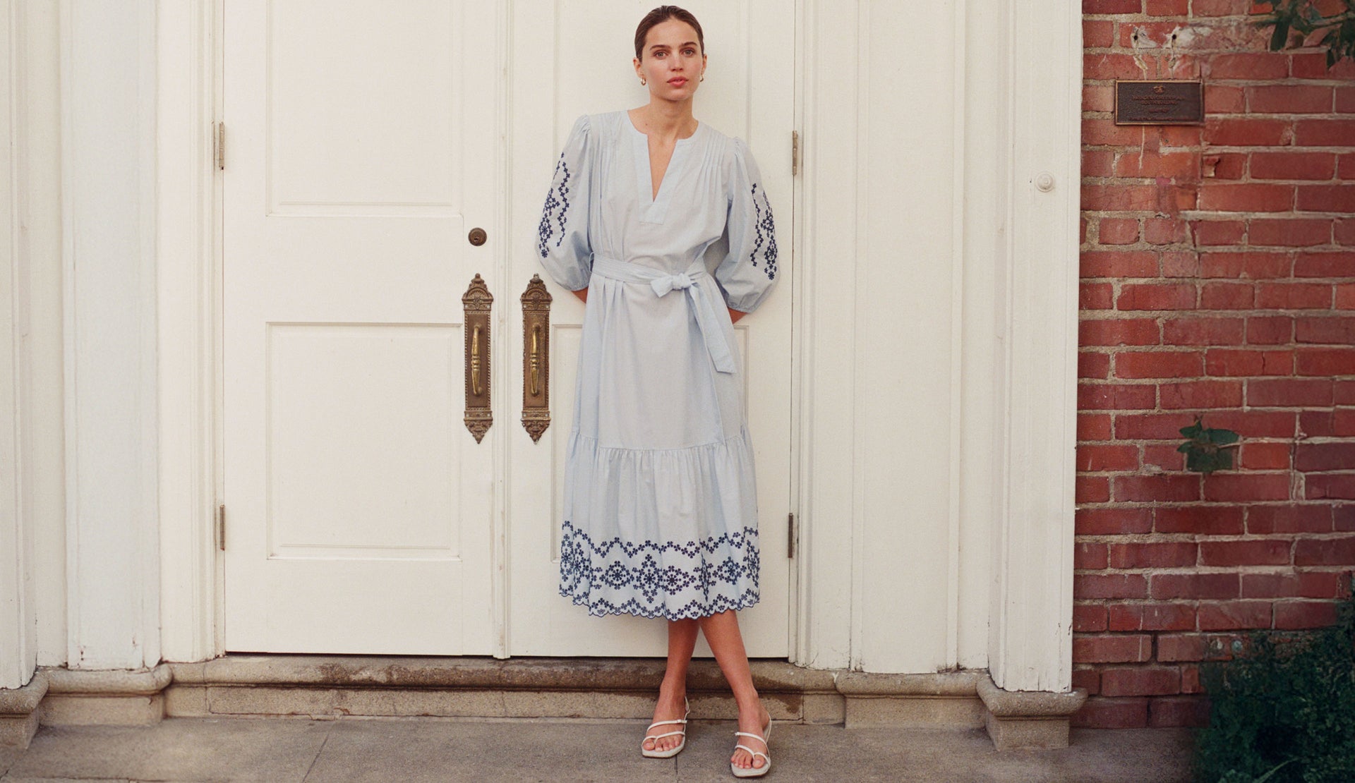 Woman in a light blue dress with floral patterns standing in front of a white door.