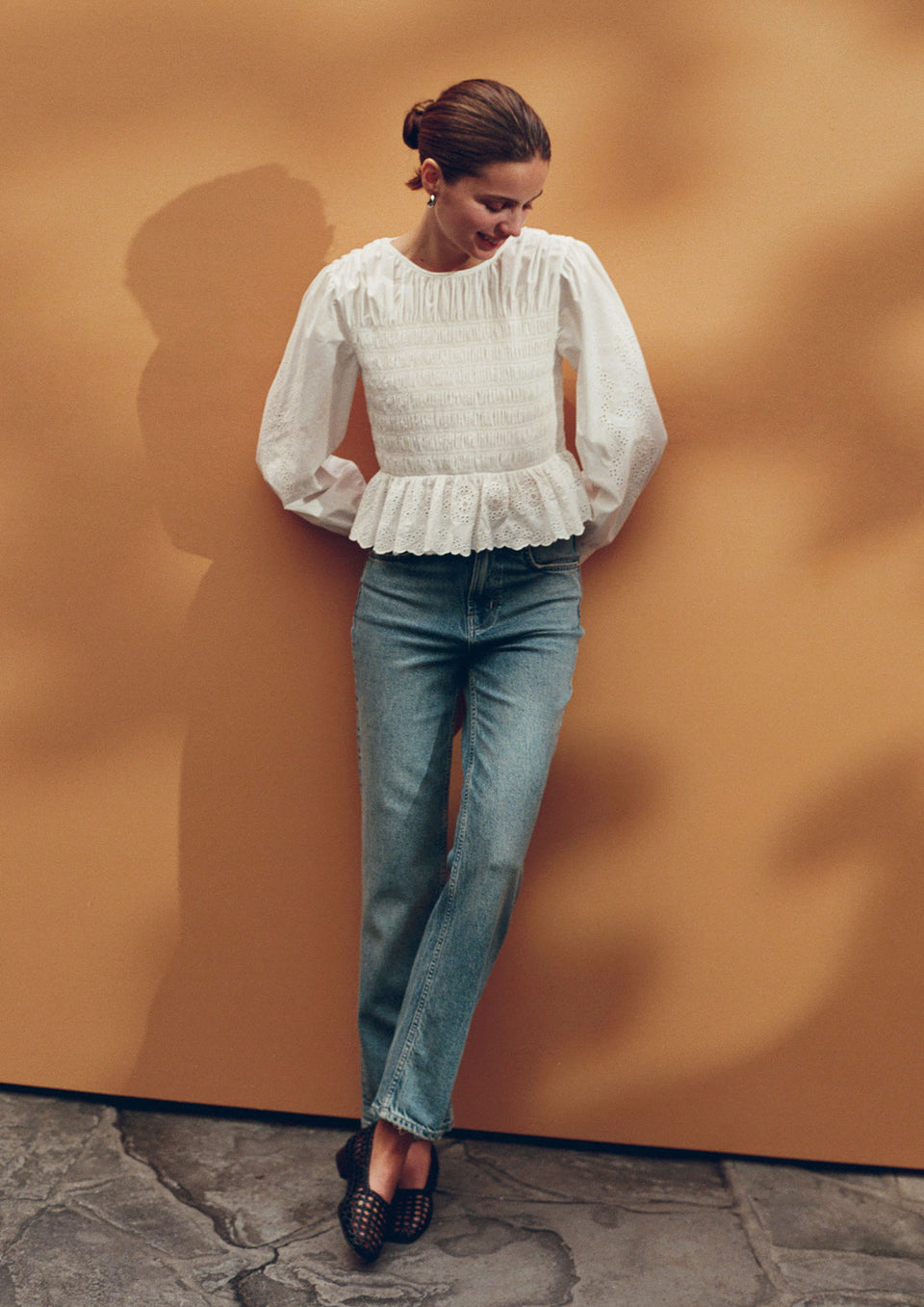 Woman wearing a white blouse with ruffled details and blue jeans against an orange wall.