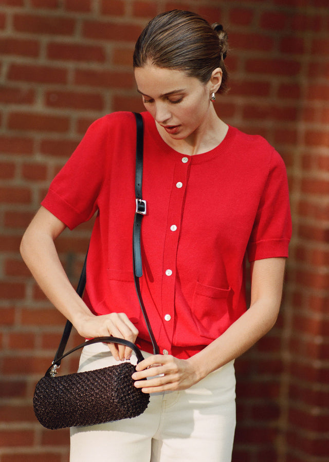 Woman in a red shirt holding a black handbag against a brick wall.