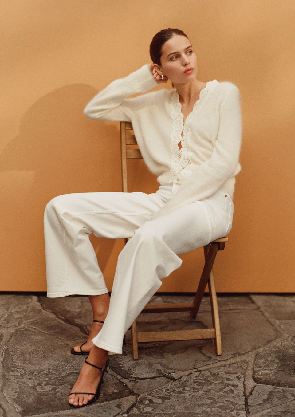 Woman in a white outfit sitting on a wooden chair against an orange wall.