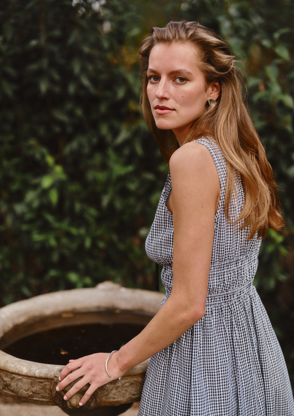 Woman in a checkered dress standing next to a stone fountain with greenery in the background