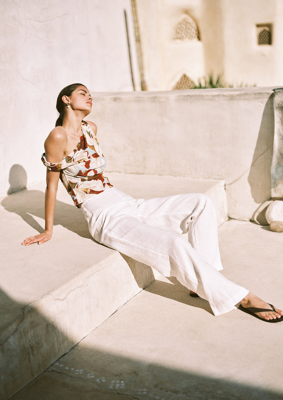 Woman in a floral top and white pants sitting on a stone bench with a neutral background