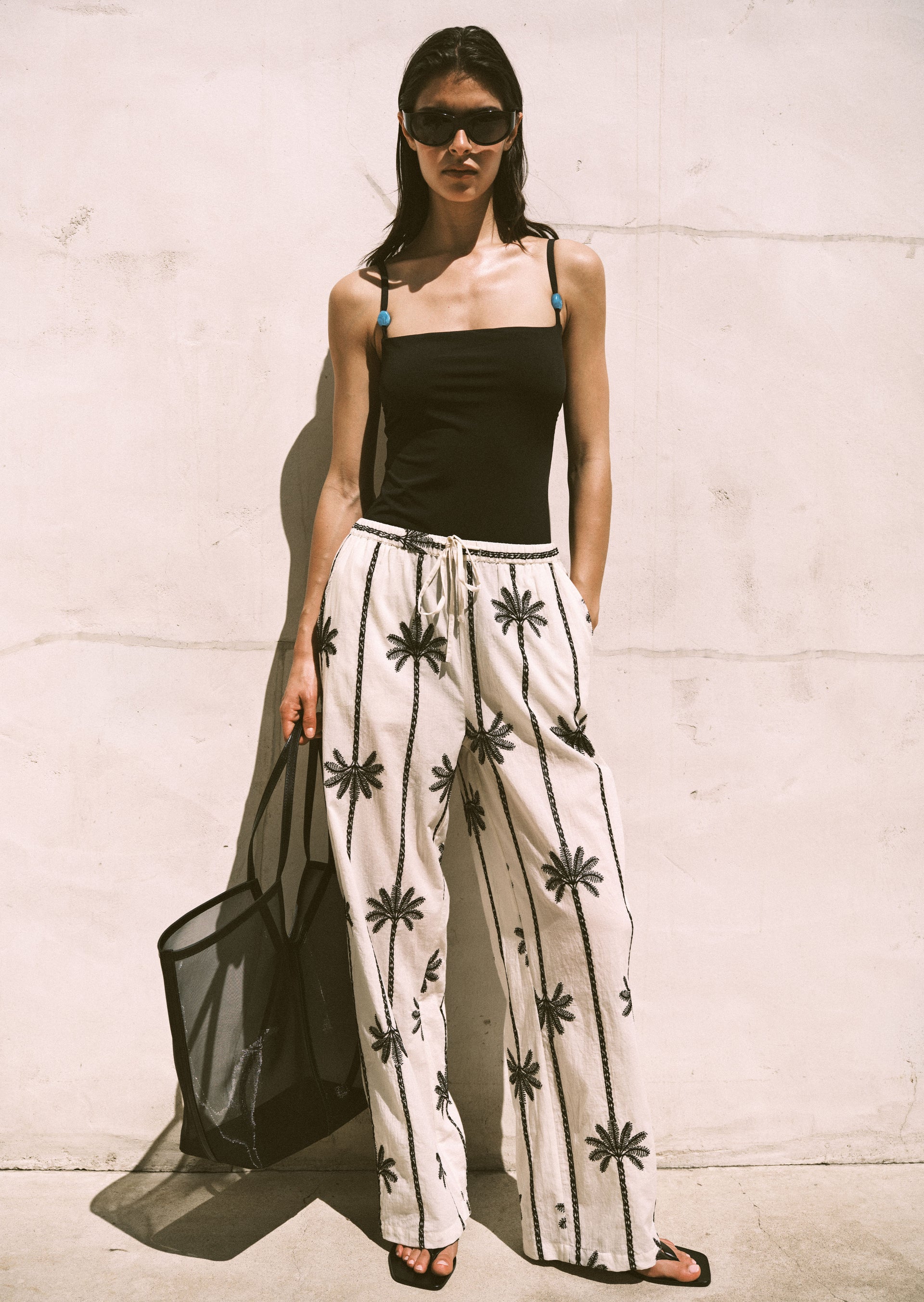 Woman wearing a black bikini and beige pants with black patterns against a light-colored wall.