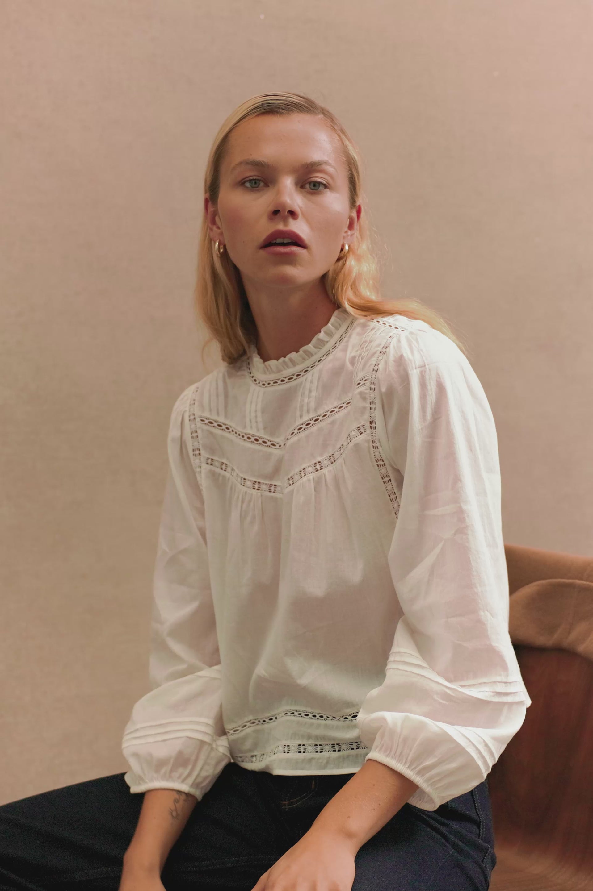 Stylized front view of female model wearing our Reyna Top in white. This is a long sleeve crewneck baby doll blouse, with ruffles at the collar. 