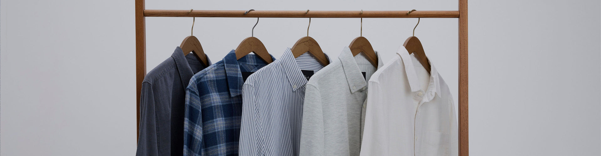 A wooden clothing rack holding five shirts in different colors and patterns, including dark gray, blue plaid, light blue, light gray, and off-white, against a plain white background.